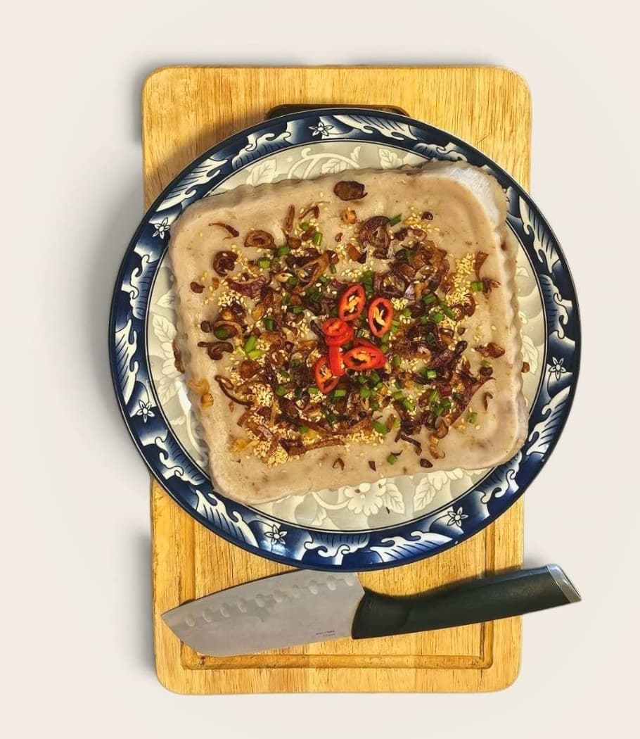 A whole traditional yam cake from Fatty Yam, on a blue and white patterned plate, viewed from above, garnished with fried shallots, spring onion, sesame and red chilli